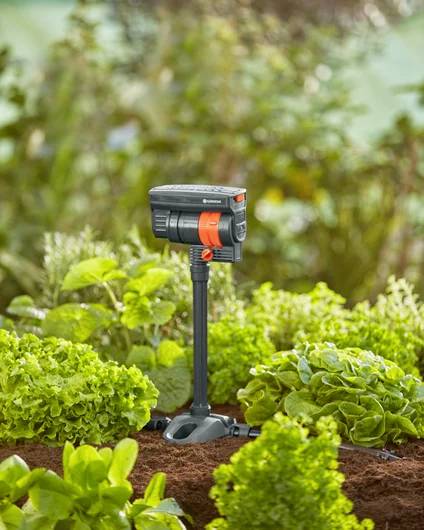 Eine kompakte Sprinkleranlage mit schwarz-orangefarbener Gestaltung ist inmitten üppiger grüner Pflanzen im Garten platziert und sorgt für Bewässerung.