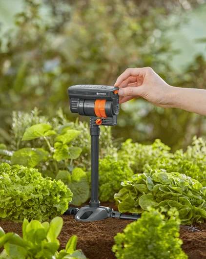 Eine Hand stellt einen Gartenbewässerungstimer zwischen üppig grünen Pflanzen ein, was die automatisierte Steuerung der Bewässerung veranschaulicht.