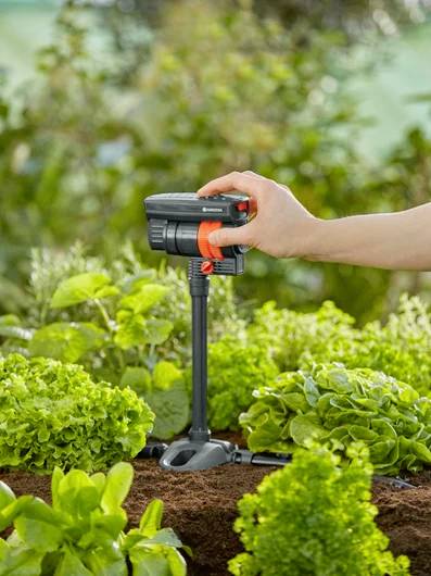Eine Hand justiert ein Gartenbewässerungssystem zwischen grünen Pflanzen und veranschaulicht die Verwendung von Bewässerungsausrüstung beim Gärtnern.