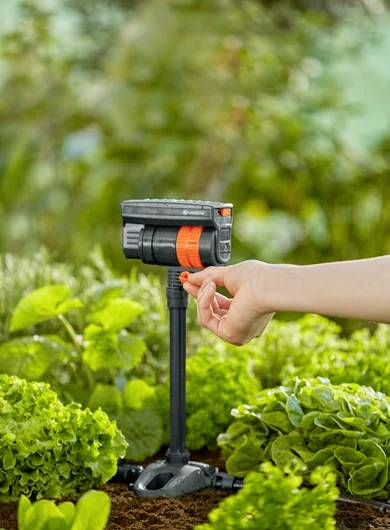 Eine Hand justiert eine schwarze und orangefarbene Wasserdüse in einem Garten, umgeben von grünen Pflanzen und Erde, was auf einen Bewässerungsaufbau hindeutet.