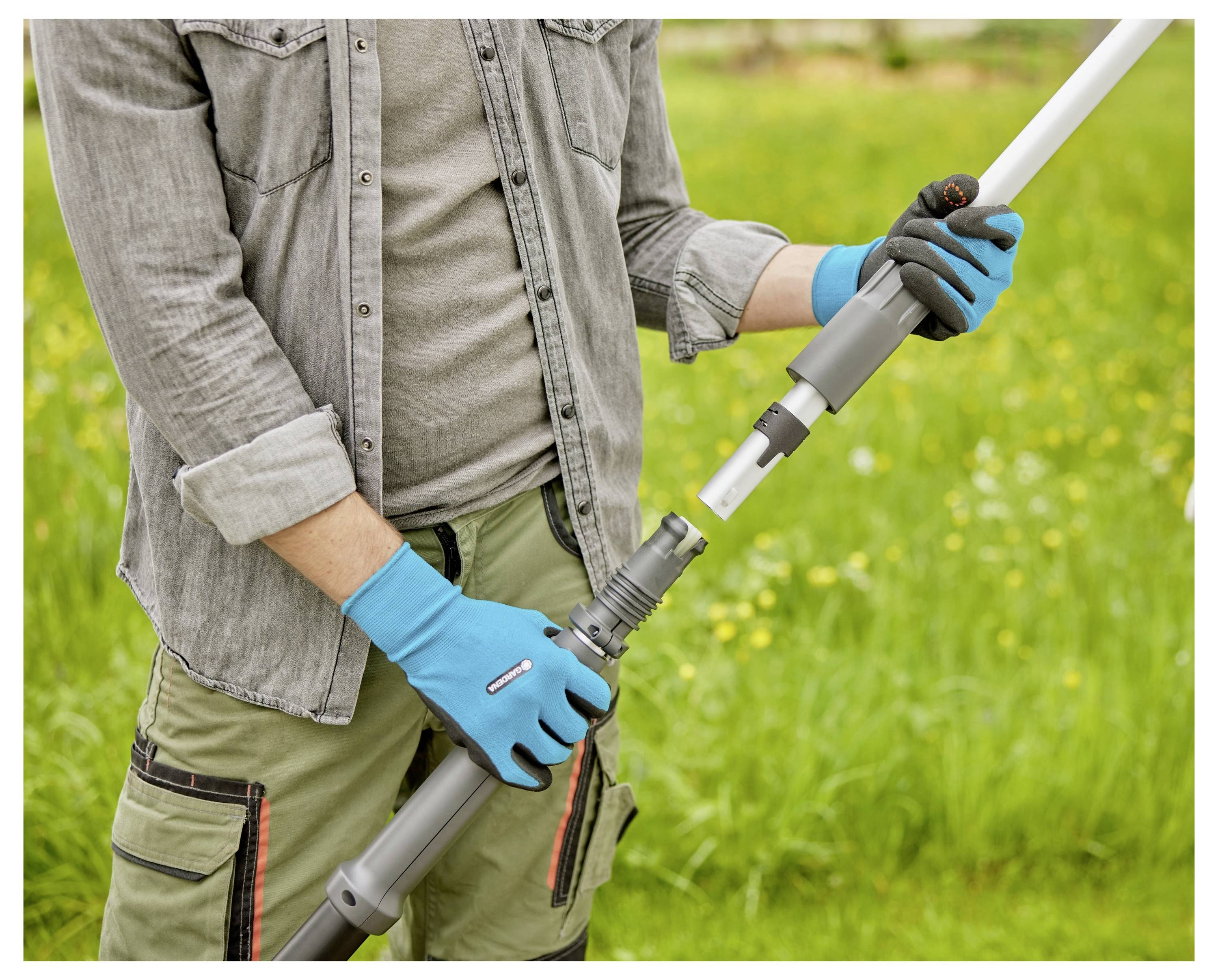Eine Person, die blaue Handschuhe trägt, verbindet zwei Abschnitte von Gartenschlauch in einer grasbewachsenen Außenumgebung und deutet damit auf Montage- oder Reparaturarbeiten hin.