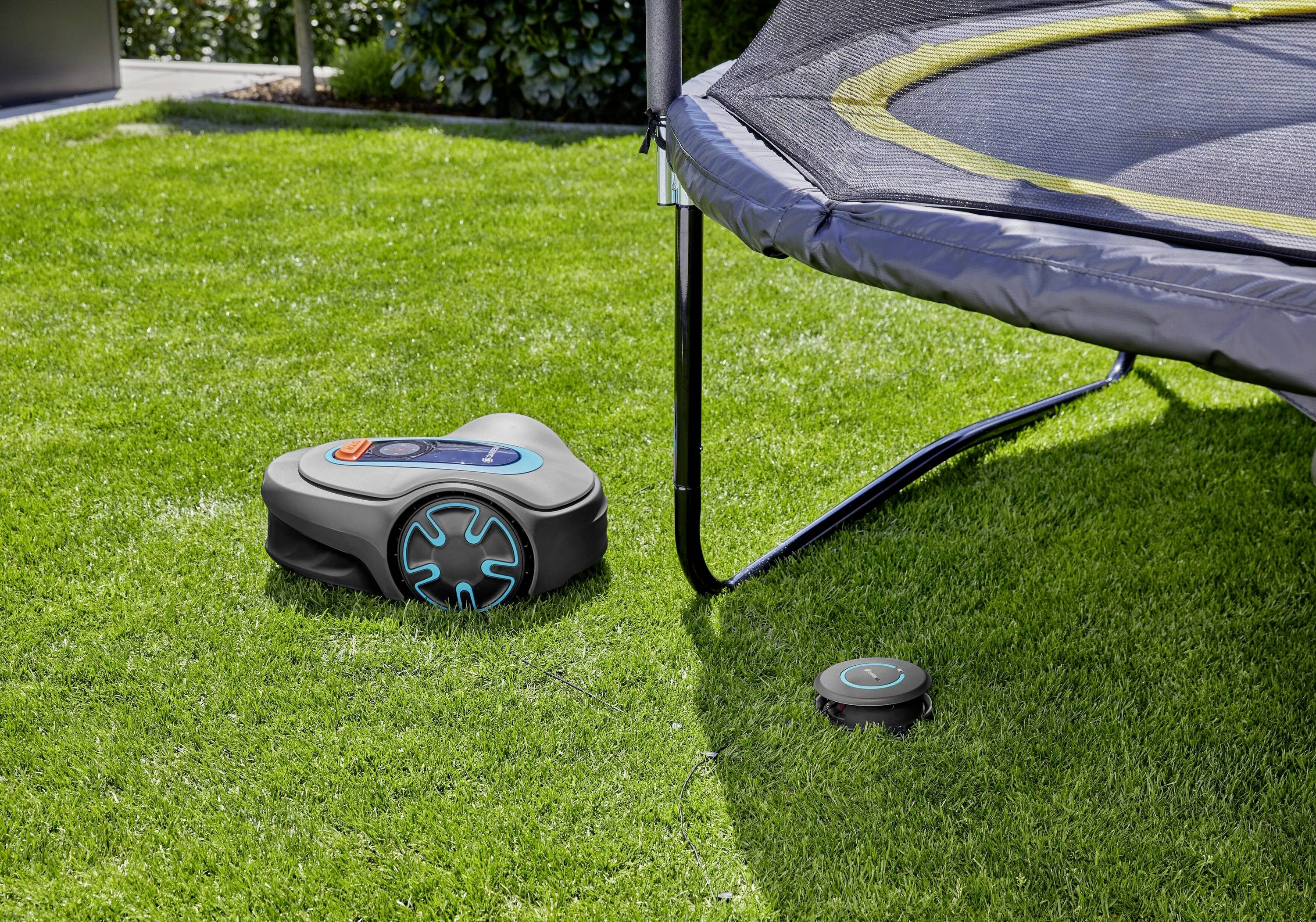 Ein Rasenroboter mäht Gras neben einem Trampolin im Garten. Ein kleines Gerät liegt auf dem Rasen als Teil des Mähsystems.