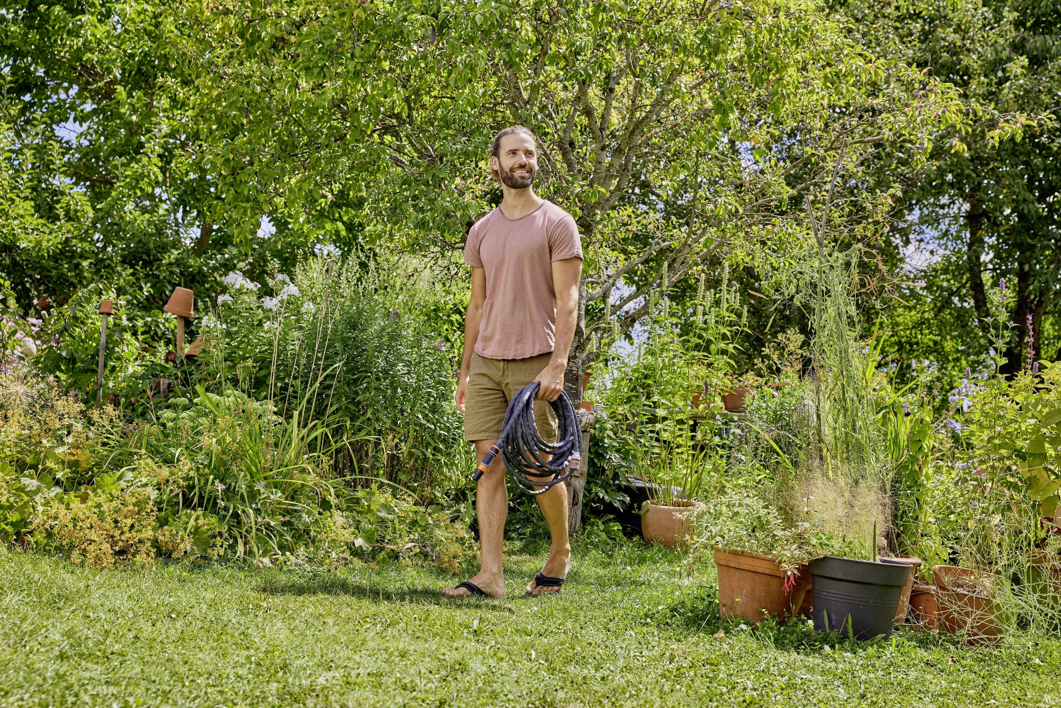 Mann mit Schlauch geht durch üppigen Garten mit vielen Pflanzen und Bäumen, sonniges Wetter, entspannte Atmosphäre.