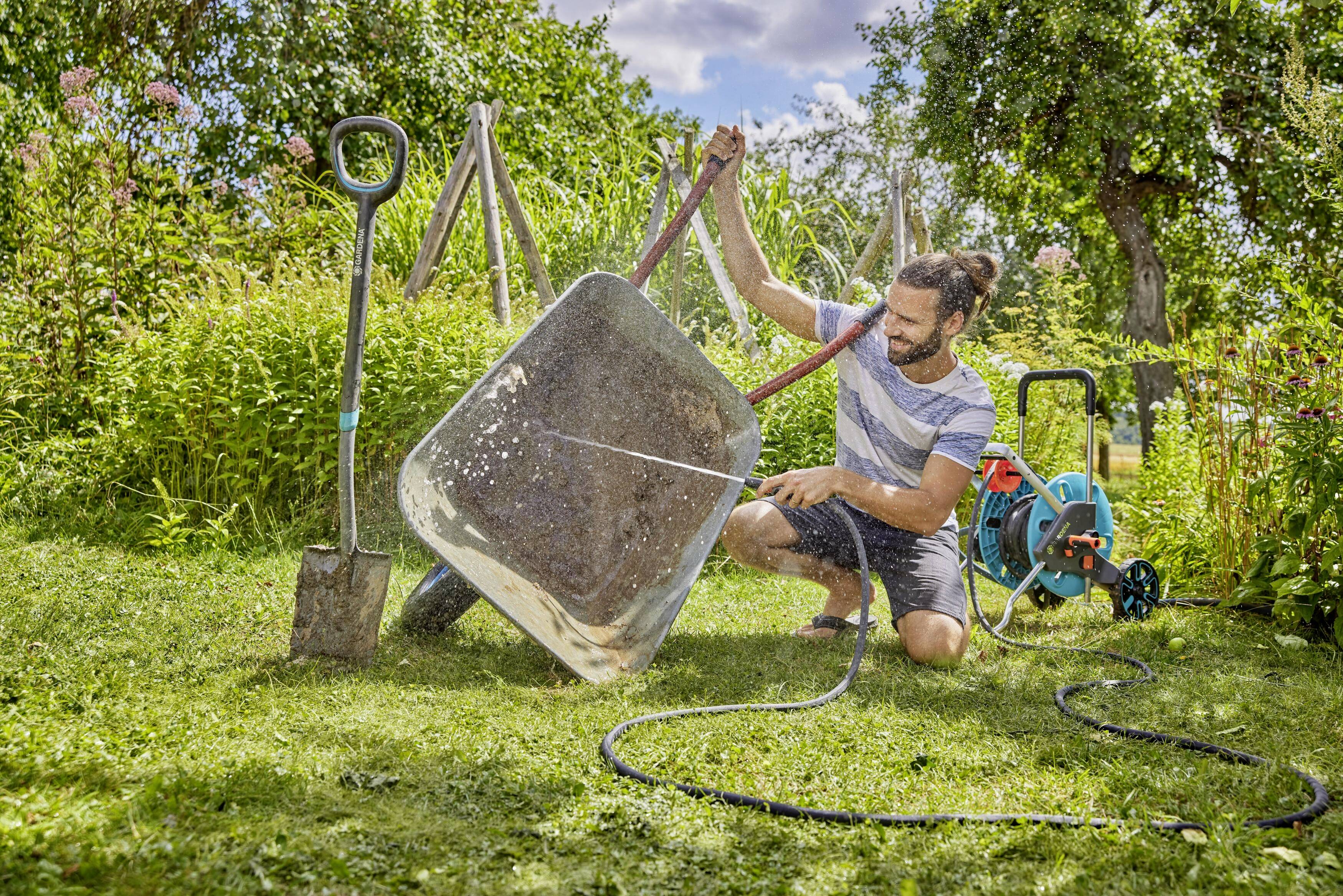 Eine Person reinigt im Garten mit einem Wasserschlauch eine Schubkarre. Daneben steht eine Schaufel. Es ist sonnig und grün.