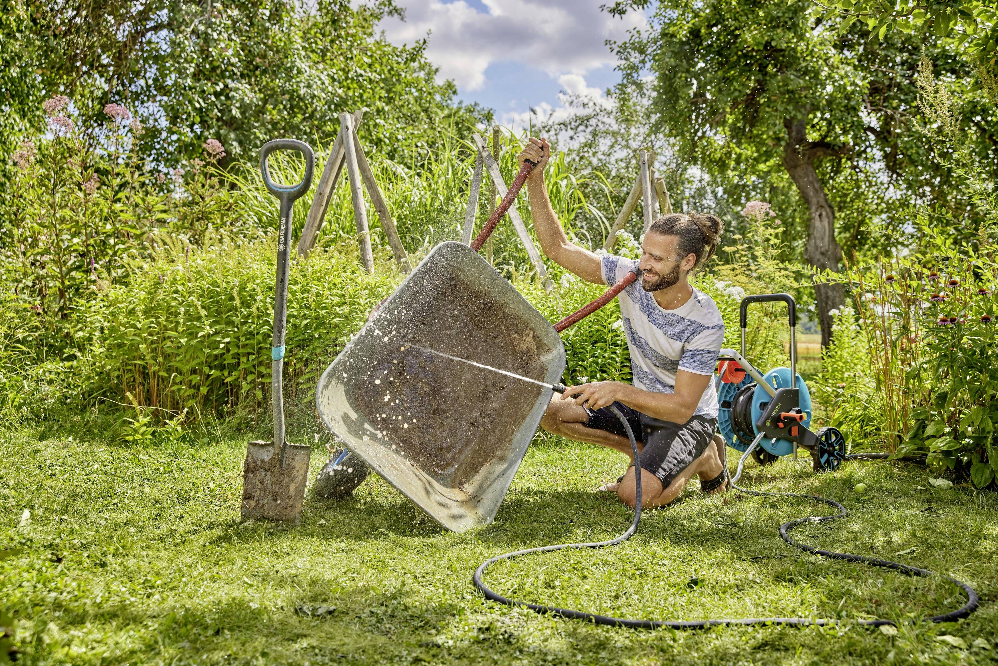Ein Mann reinigt eine Schubkarre mit einem Gartenschlauch im Garten, umgeben von Bäumen und Pflanzen.