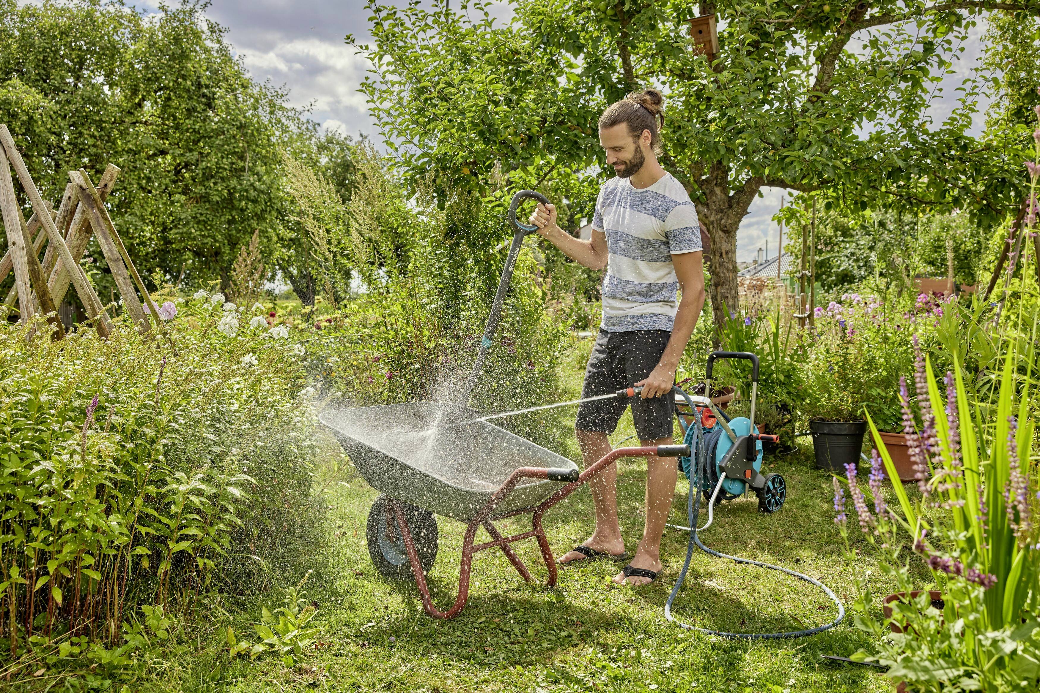 Ein Mann bewässert eine Schubkarre im Garten mit einem Schlauch, umgeben von Pflanzen und Bäumen bei sonnigem Wetter.
