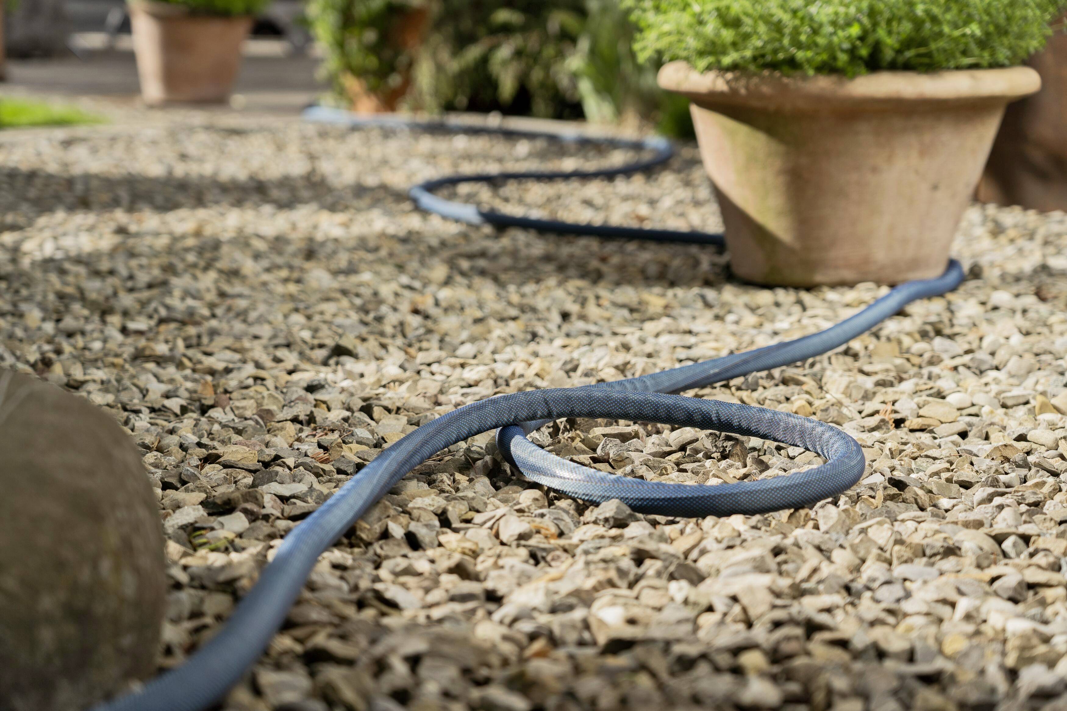 Ein Wasserschlauch verläuft gewunden über einen Kiesweg zwischen zwei Topfpflanzen im Garten.