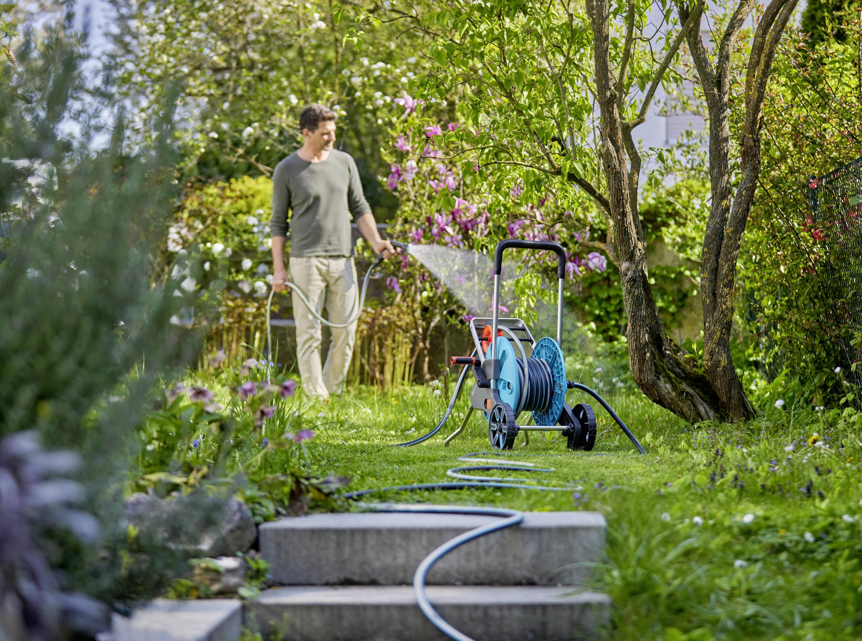Ein Mann bewässert einen Garten mit einem Gartenschlauch neben blühenden Büschen und einem Baum. Im Vordergrund ist eine Schlauchtrommel sichtbar.