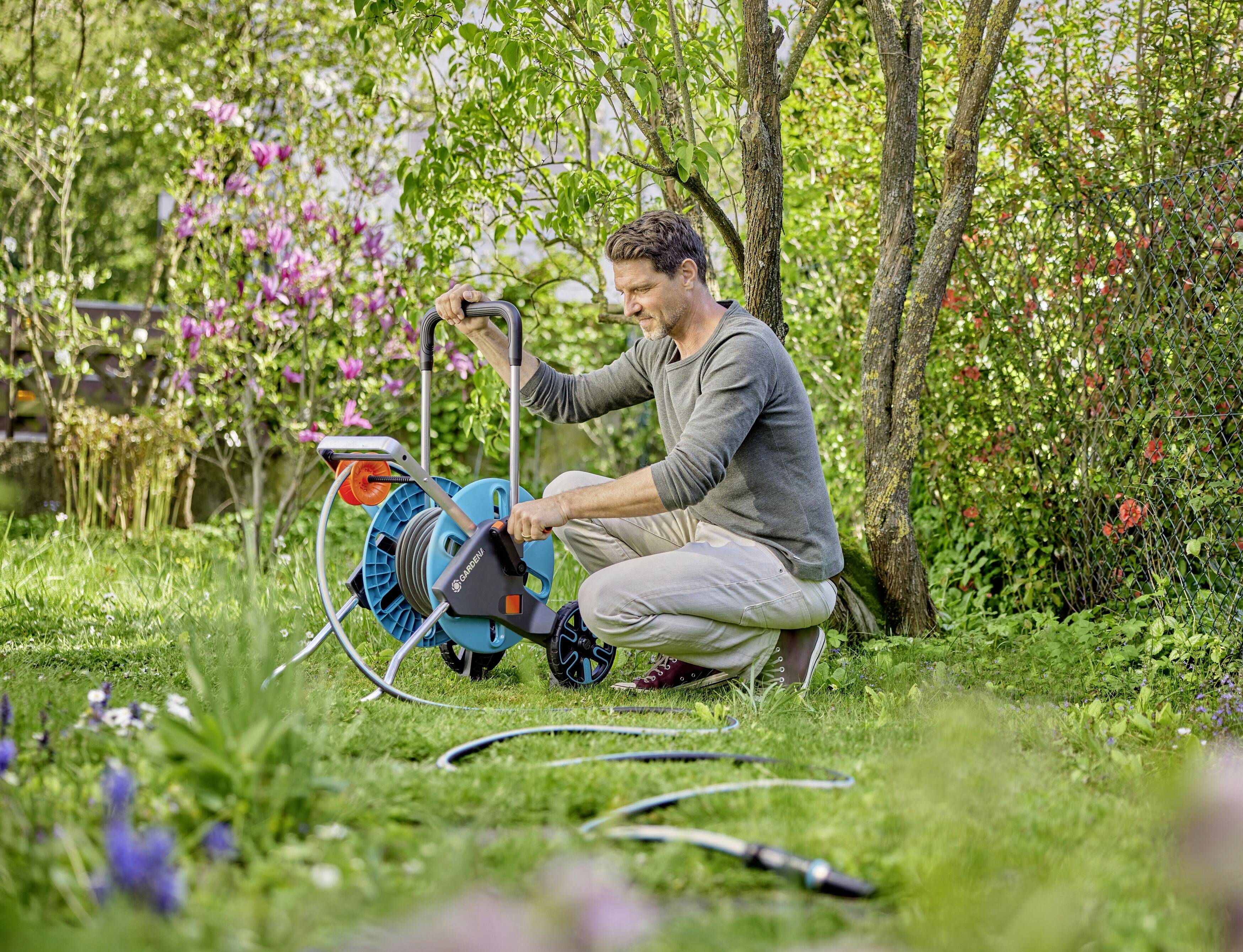 Eine Person im Garten dreht den Schlauch einer Schlauchtrommel auf. Im Hintergrund sind grüne Pflanzen und blühende Büsche zu sehen.
