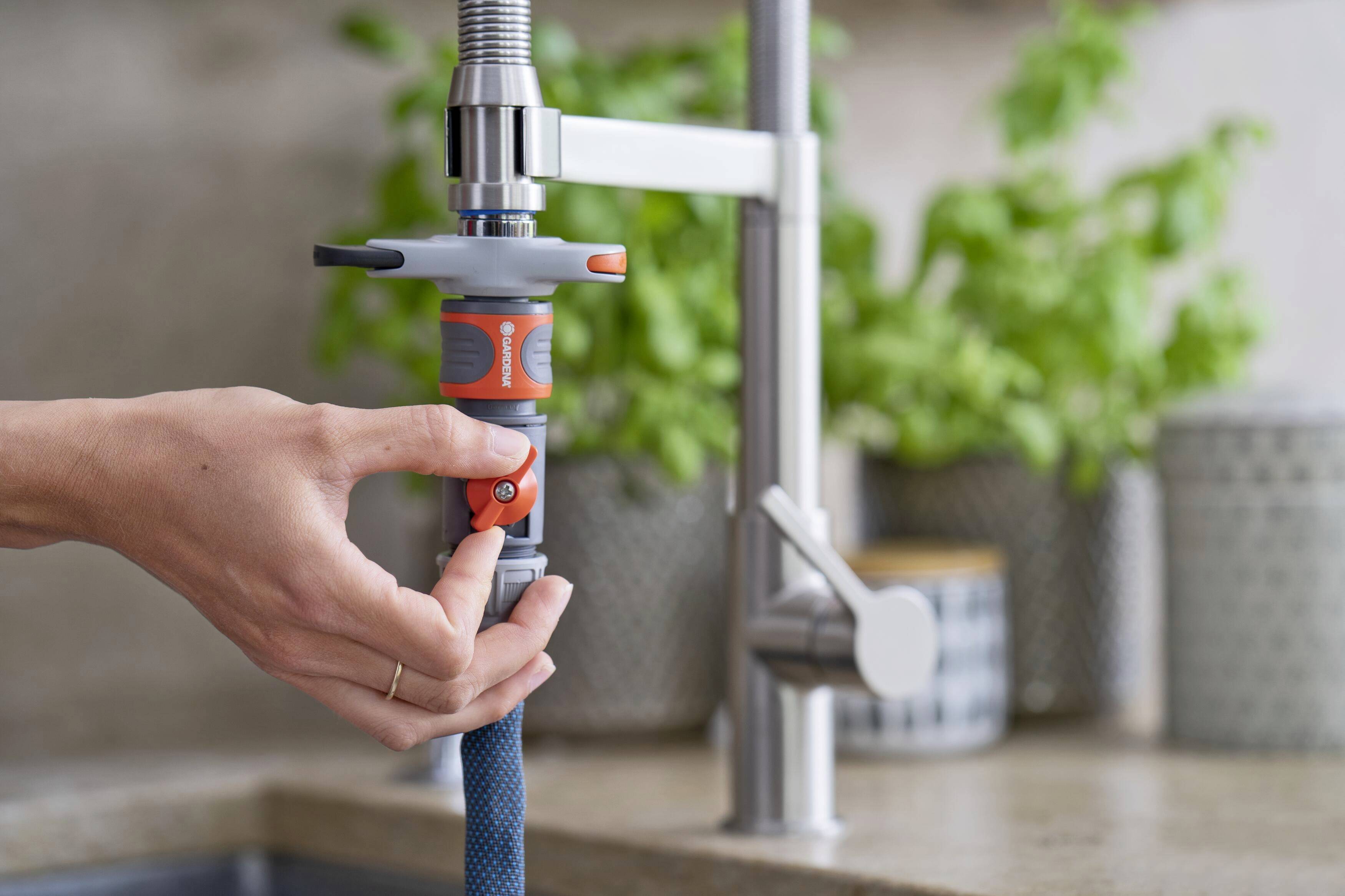 Eine Hand befestigt einen Wasserschlauch an einem Küchenarmaturadapter. Im Hintergrund sind grüne Pflanzen auf der Arbeitsplatte zu sehen.