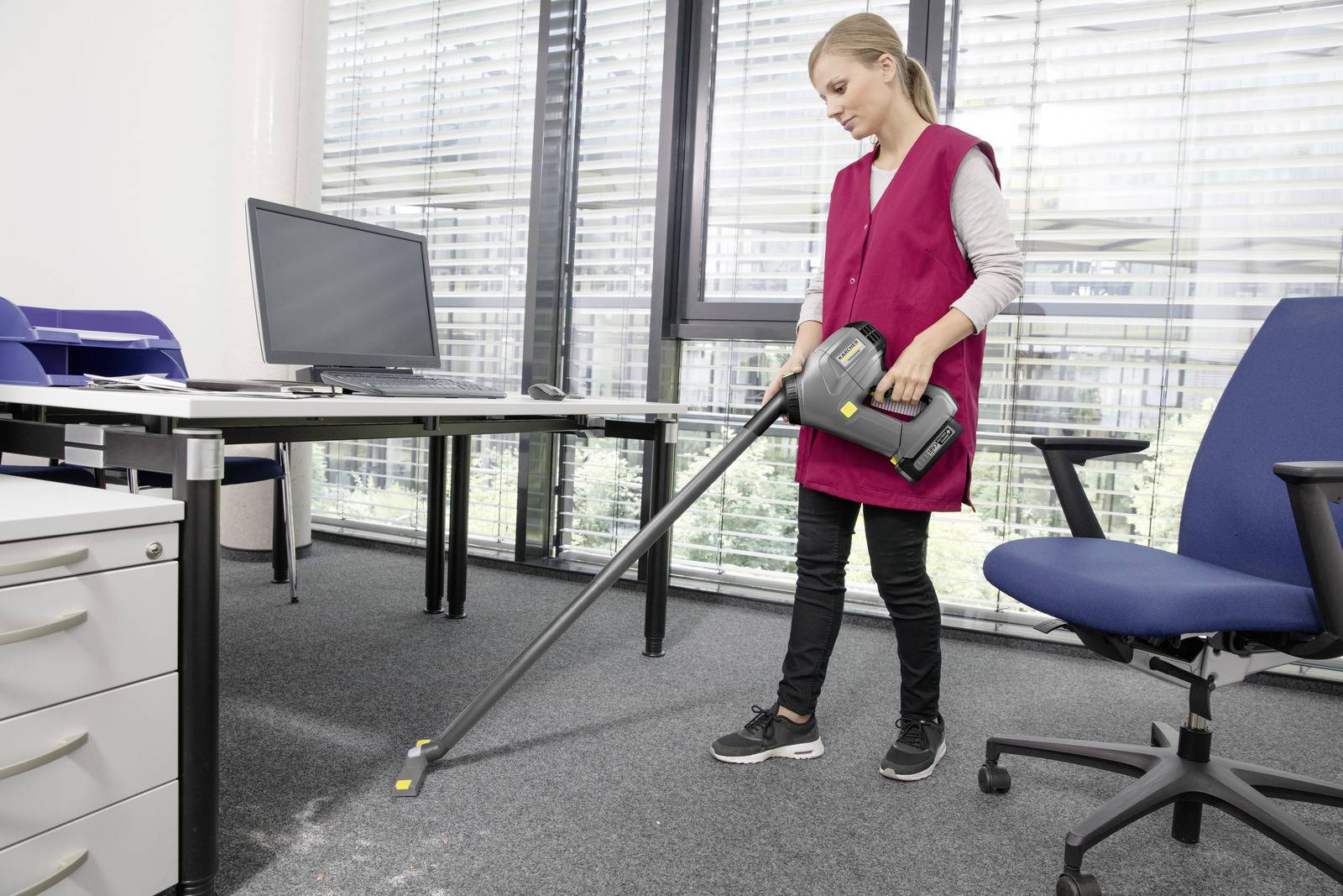 Eine Person in einer rosa Weste saugt einen Teppichboden in einem Büro in der Nähe eines Schreibtisches mit einem Computermonitor, Aktenschränken und einem Bürostuhl.