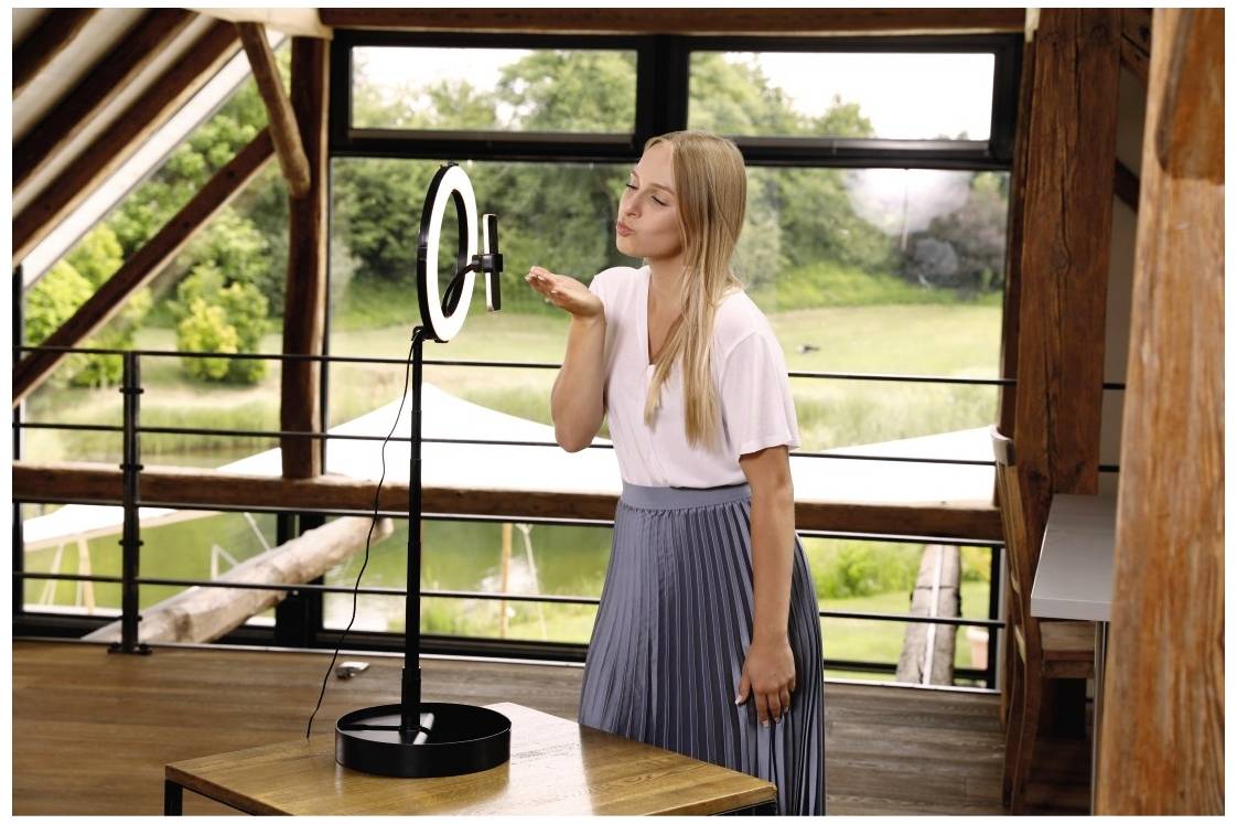 Frau in hellem Raum, stehend vor Ringlicht und Kamera, wirkt als ob sie in Kamera küsst. Fenster im Hintergrund mit grünem Ausblick.