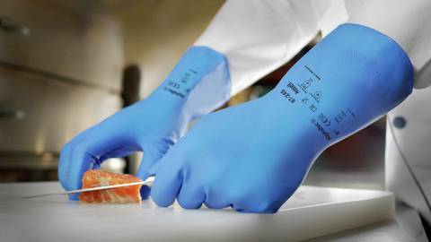 Eine Person in blauen Handschuhen schneidet Lachs mit einem Messer auf einem Schneidebrett in einer Küche.