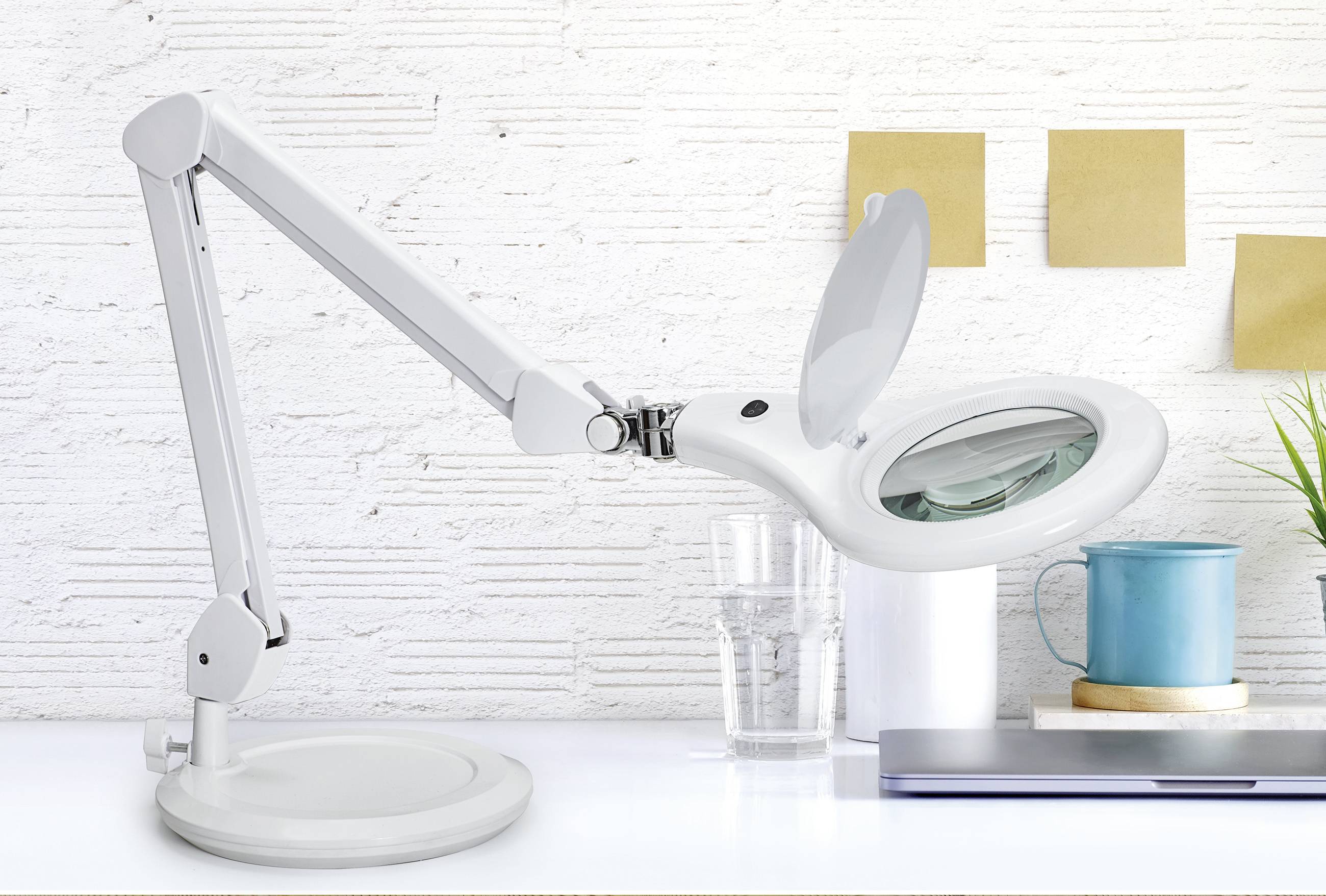 Eine weiße Schreibtischlampe mit Vergrößerungsglas steht auf einem Tisch. Im Hintergrund sind Notizen, ein Glas, eine Tasse und eine Laptop-Tastatur zu sehen.