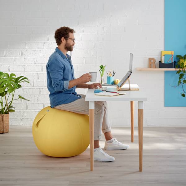 Ein Mann arbeitet an einem Schreibtisch, sitzt auf einem gelben Gymnastikball, umgeben von Büropflanzen und moderner Dekoration.