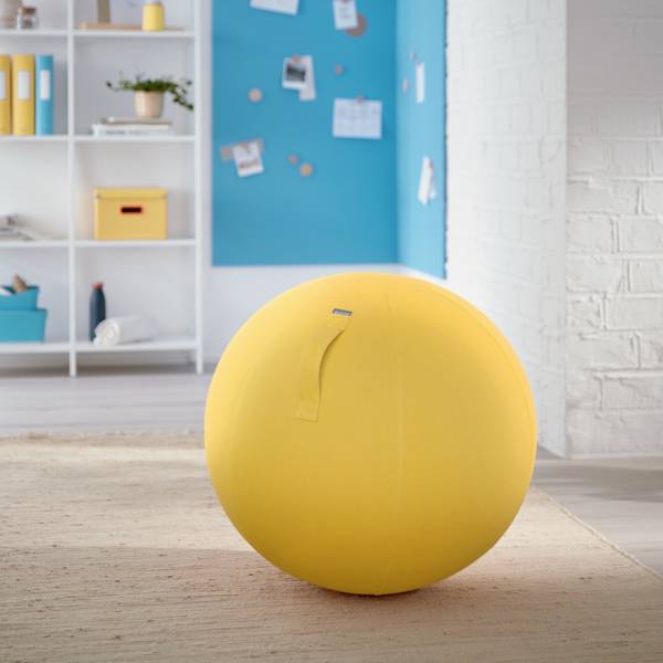 Ein großer gelber Gymnastikball steht in einem hellen Raum vor einem blau-weißen Hintergrund mit Regalen und Pinnwand.