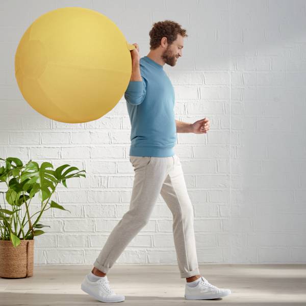 Ein Mann in blauem Pullover hält einen großen gelben Gymnastikball. Daneben steht eine Pflanze in einem Topf vor einer weißen Ziegelwand.