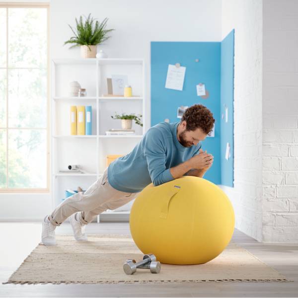 Mann trainiert mit großem gelben Gymnastikball im hellen Raum; Hanteln liegen auf Teppich.