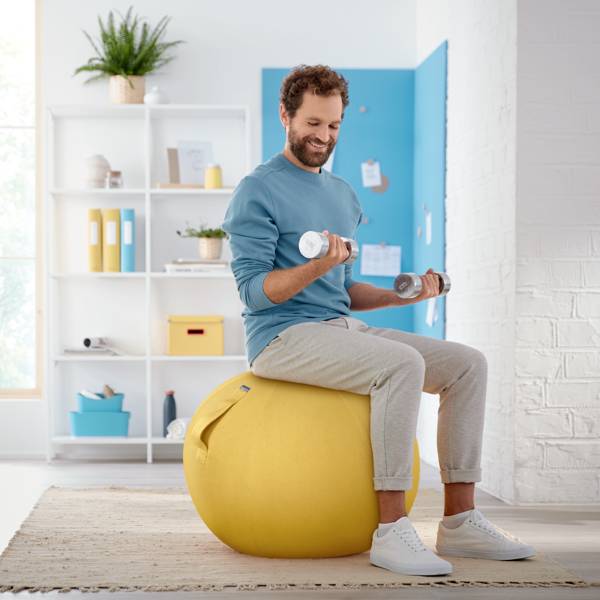 Ein Mann trainiert mit Hanteln auf einem gelben Ball im Wohnzimmer. Im Hintergrund sind ein Regal mit Büchern und Pflanzen zu sehen.