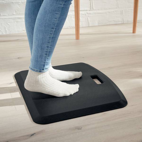 Eine Person in blauen Jeans und weißen Socken steht auf einem schwarzen Balance-Board auf einem Holzboden.