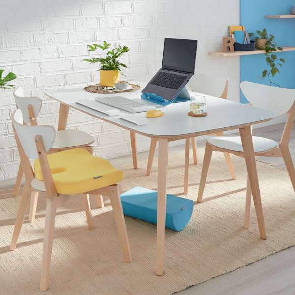 Ein moderner, heller Raum mit einem Tisch, Stühlen, Laptop und Pflanzen. Eine gelbe Kinderschale auf einem Stuhl.