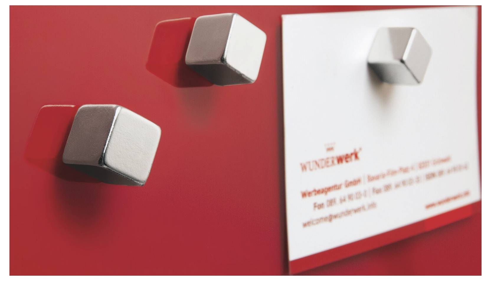 Nahaufnahme von zwei quadratischen Metallknöpfen auf einer roten Oberfläche neben einem weißen „WUNDERwerk" Etikett oder Schild.