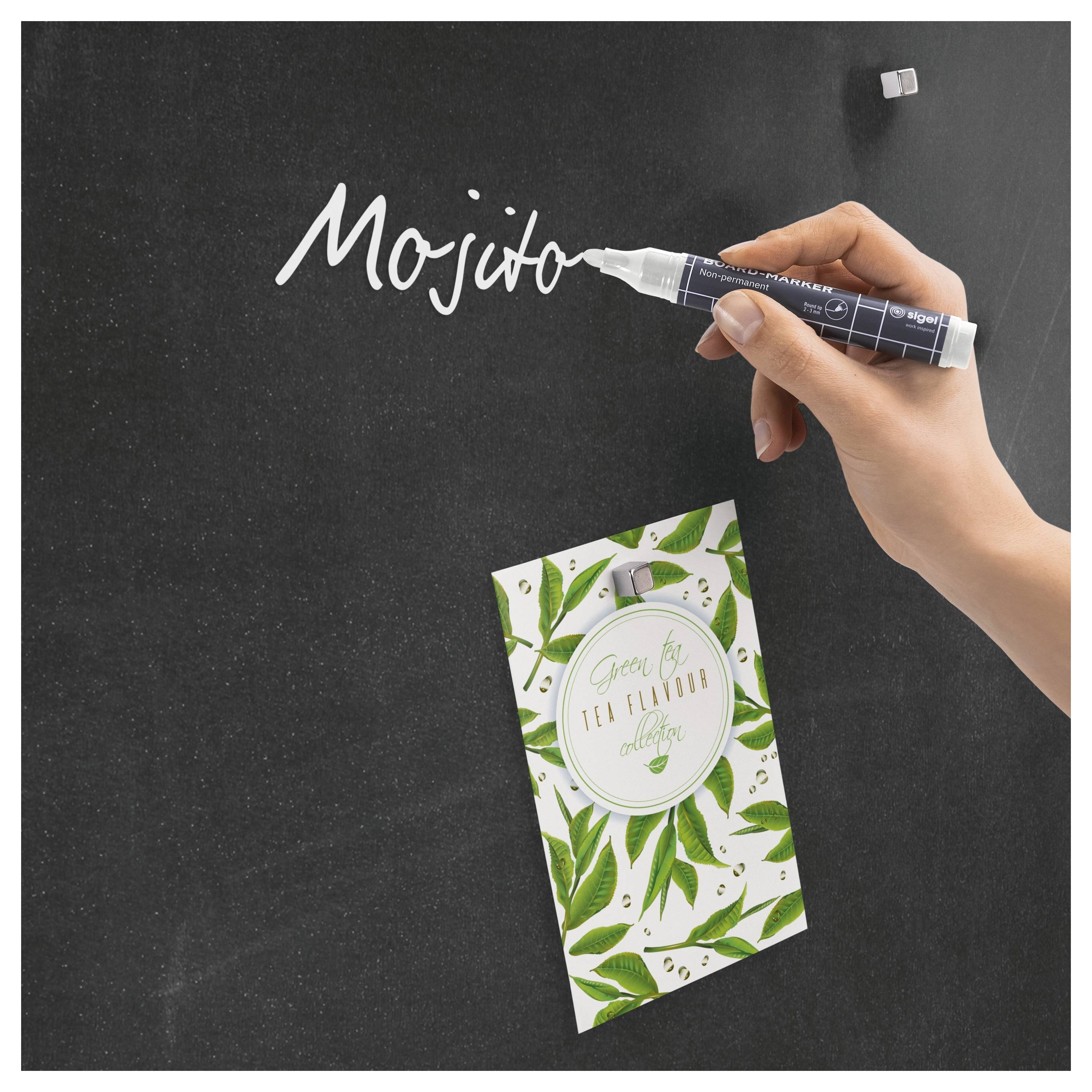 Die Hand schreibt mit einem weißen Marker das Wort „Mojito" in Weiß auf eine schwarze Oberfläche; darunter liegt eine grüne Teeverpackung mit Blattmuster.