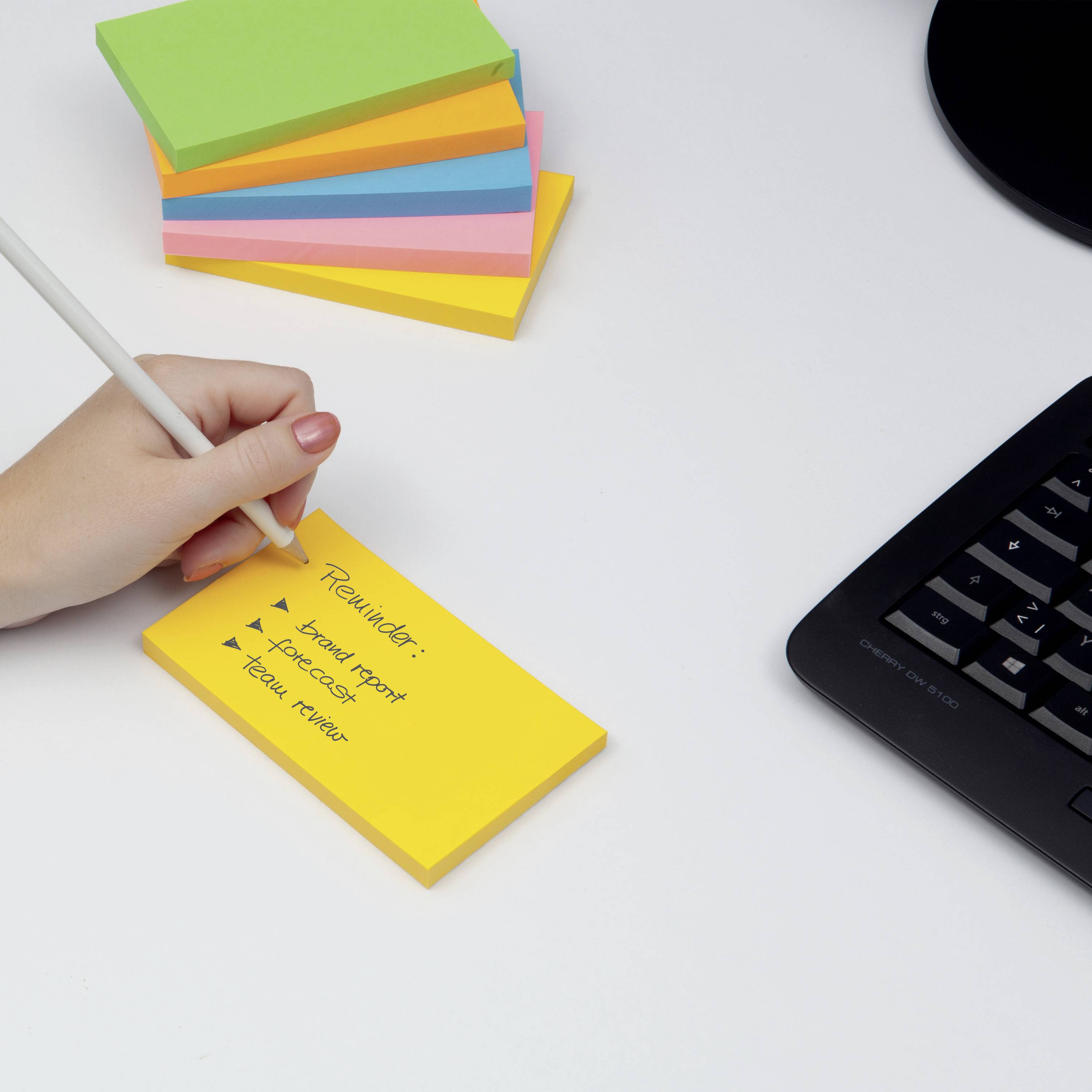 Eine Hand schreibt mit einem Stift auf ein gelbes Haftnotizpapier neben einem Computerkeyboard. Im Hintergrund sind bunte Notizzettel gestapelt.