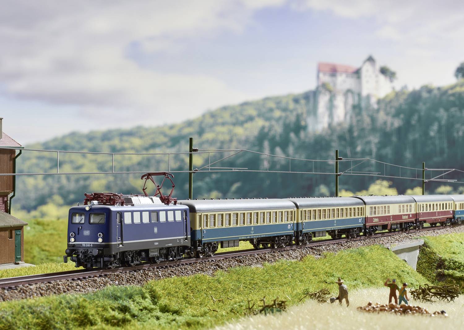 Ein Modellzug fährt durch eine ländliche Landschaft mit einem großen Schloss im Hintergrund und zwei Personen beobachten den Zug.