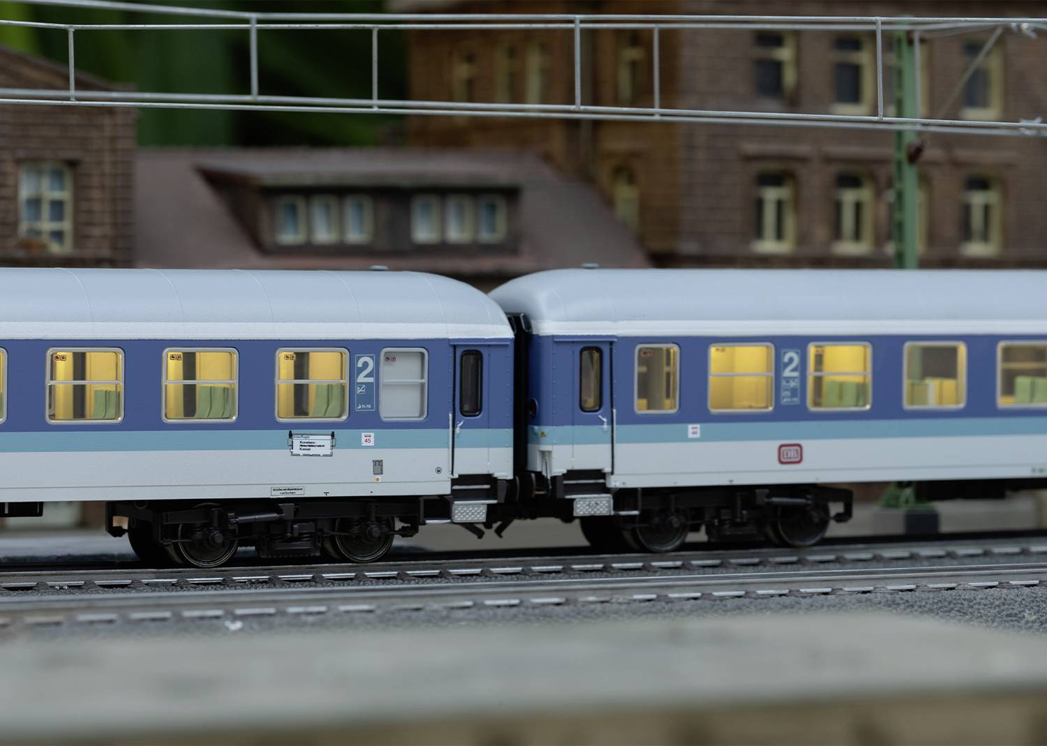 Modelleisenbahnzug mit zwei blauen, beleuchteten Waggons auf einem Gleis vor unscharfen Gebäuden im Hintergrund.