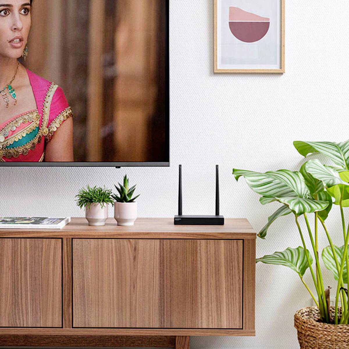 Ein modernes Wohnzimmer mit einem Holzschrank, darauf Zimmerpflanzen, ein Router und ein Flachbildfernseher, der eine Frau zeigt.