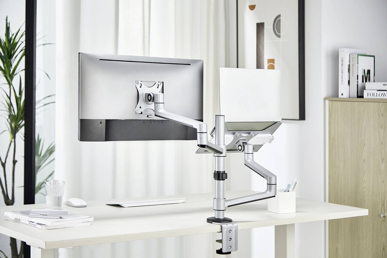 Ein Schreibtisch mit Monitorhalterung, die einen Computerbildschirm und einen Laptop nebeneinander hält. Im Hintergrund sind Pflanzen und Bilder.