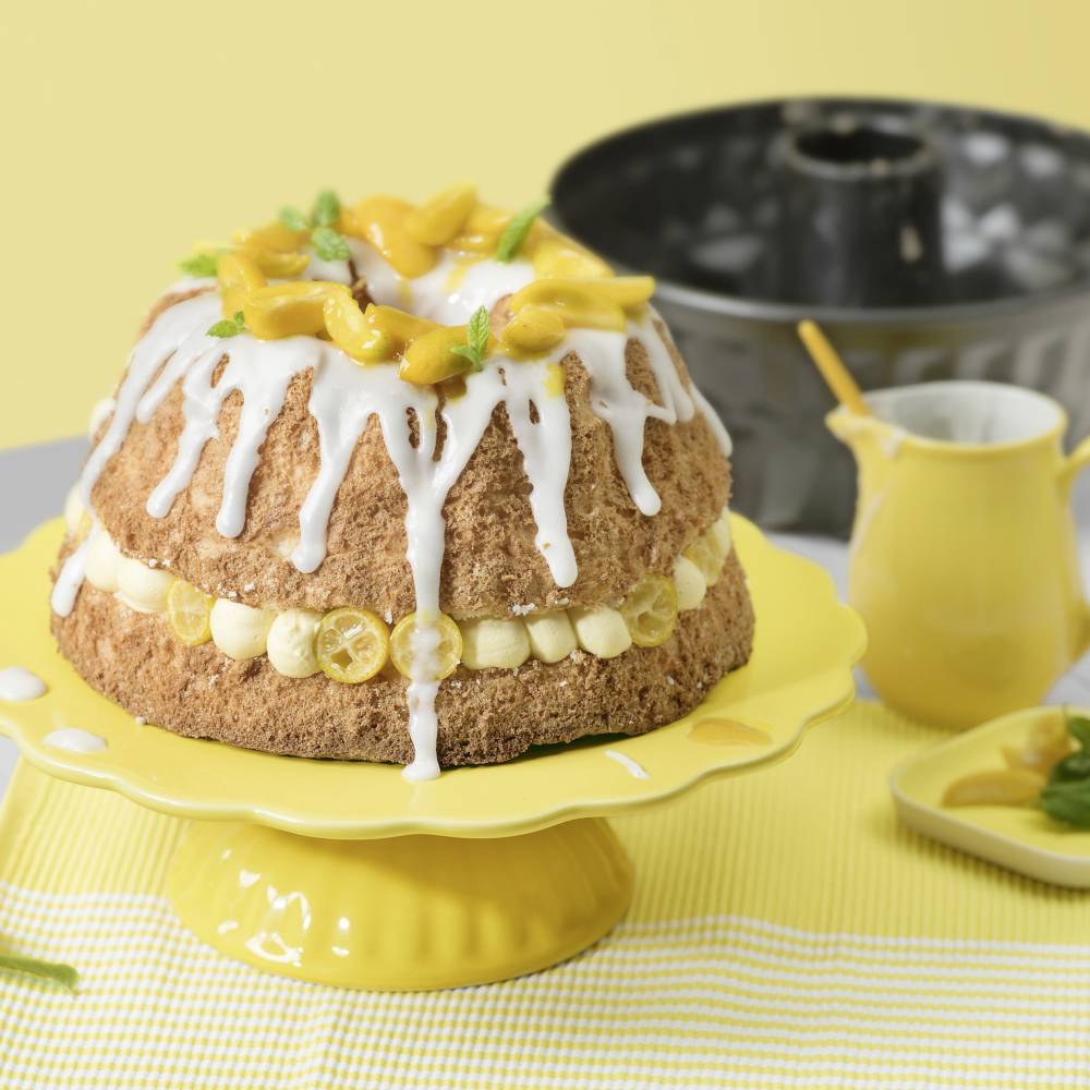 Ein Kuchen auf einem gelben Ständer, dekoriert mit Zuckerguss und Zitronenscheiben; im Hintergrund eine Kanne und eine Backform.