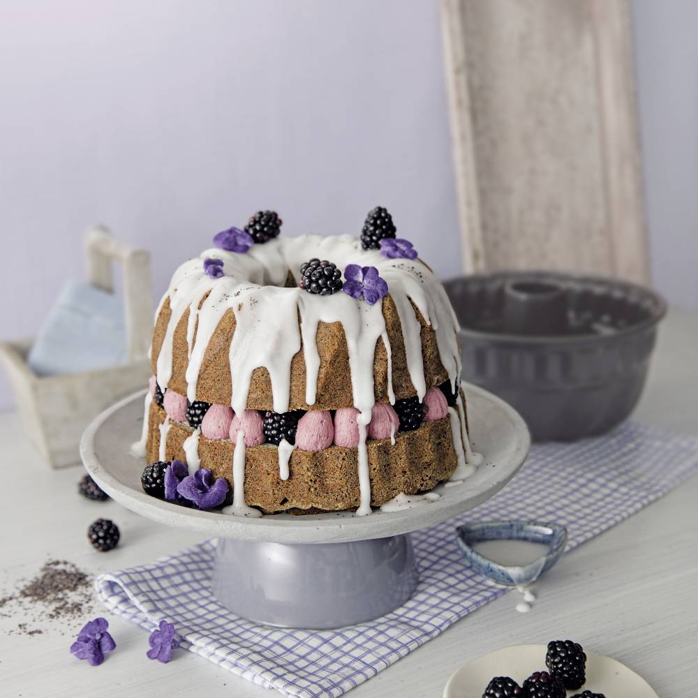 Ein dekorativer Bundt-Kuchen mit weißem Zuckerguss, dekoriert mit Brombeeren und violetten Blumen, steht auf einem Tisch. Im Hintergrund ist eine Gugelhupfform.