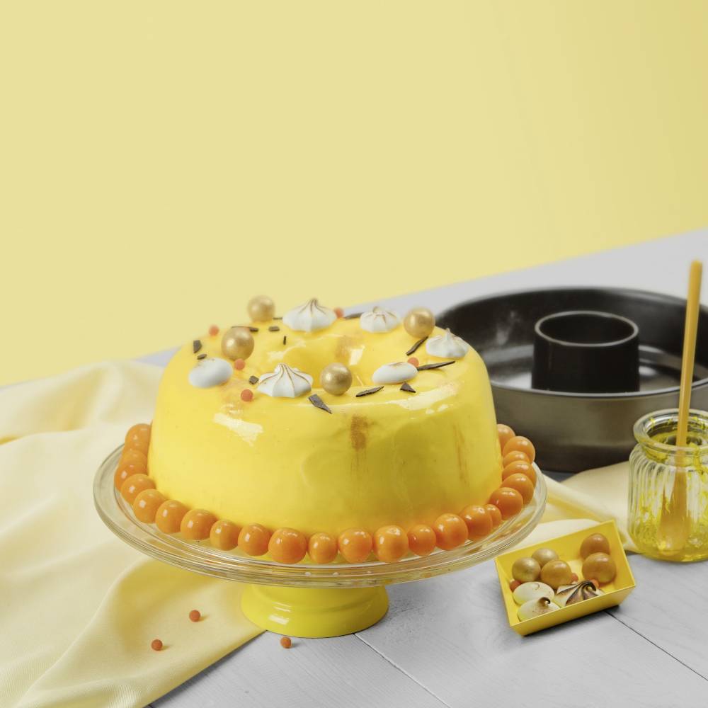 Ein gelber Kuchen mit glänzendem Überzug, dekoriert mit goldenen Kugeln und weißem Schaum, steht auf einem gelben Kuchenständer.