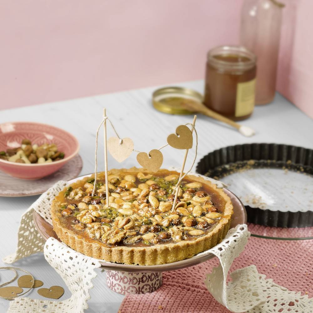 Eine mit Nüssen und Samen dekorierte Tarte steht auf einem Tisch, umgeben von leeren Tellern und einem Glas Honig im Hintergrund.