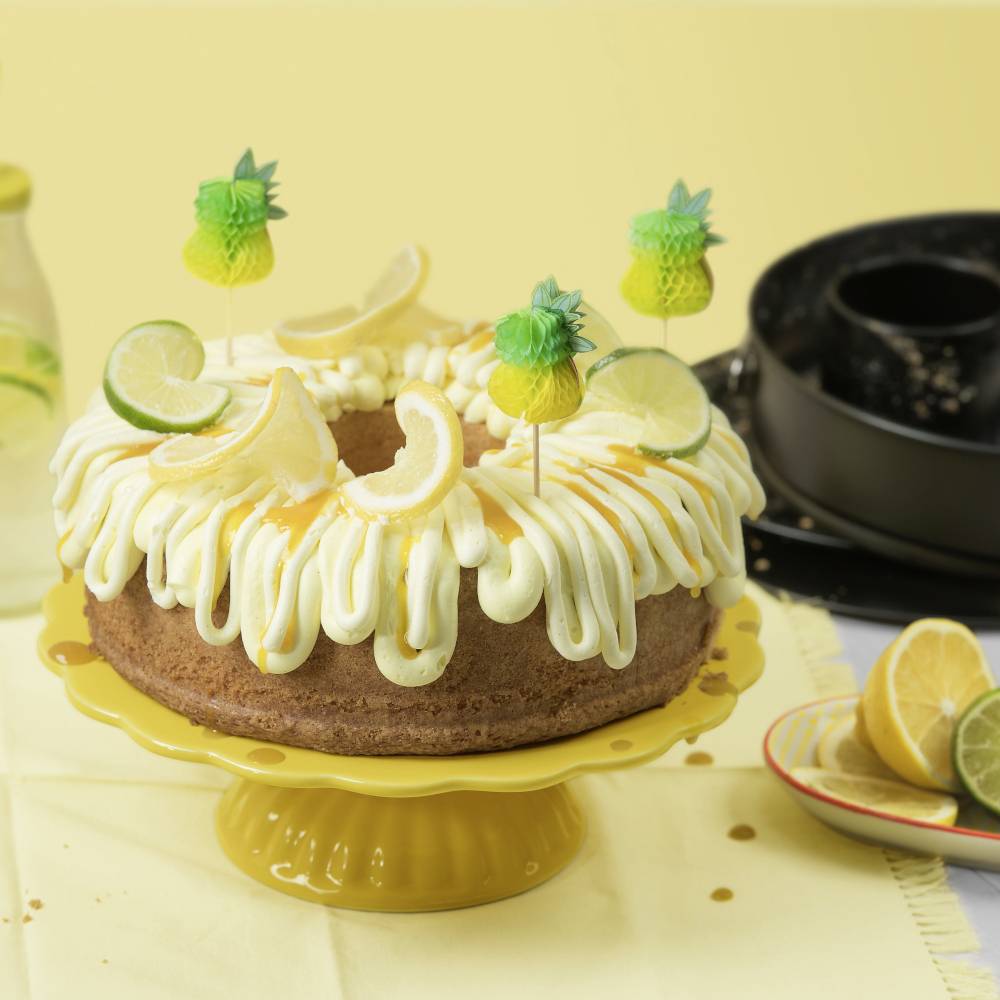Kuchen mit Zuckerguss und Zitronenscheiben, dekoriert mit Ananas-Stäbchen, auf gelbem Teller; im Hintergrund Zitronenlimonade.