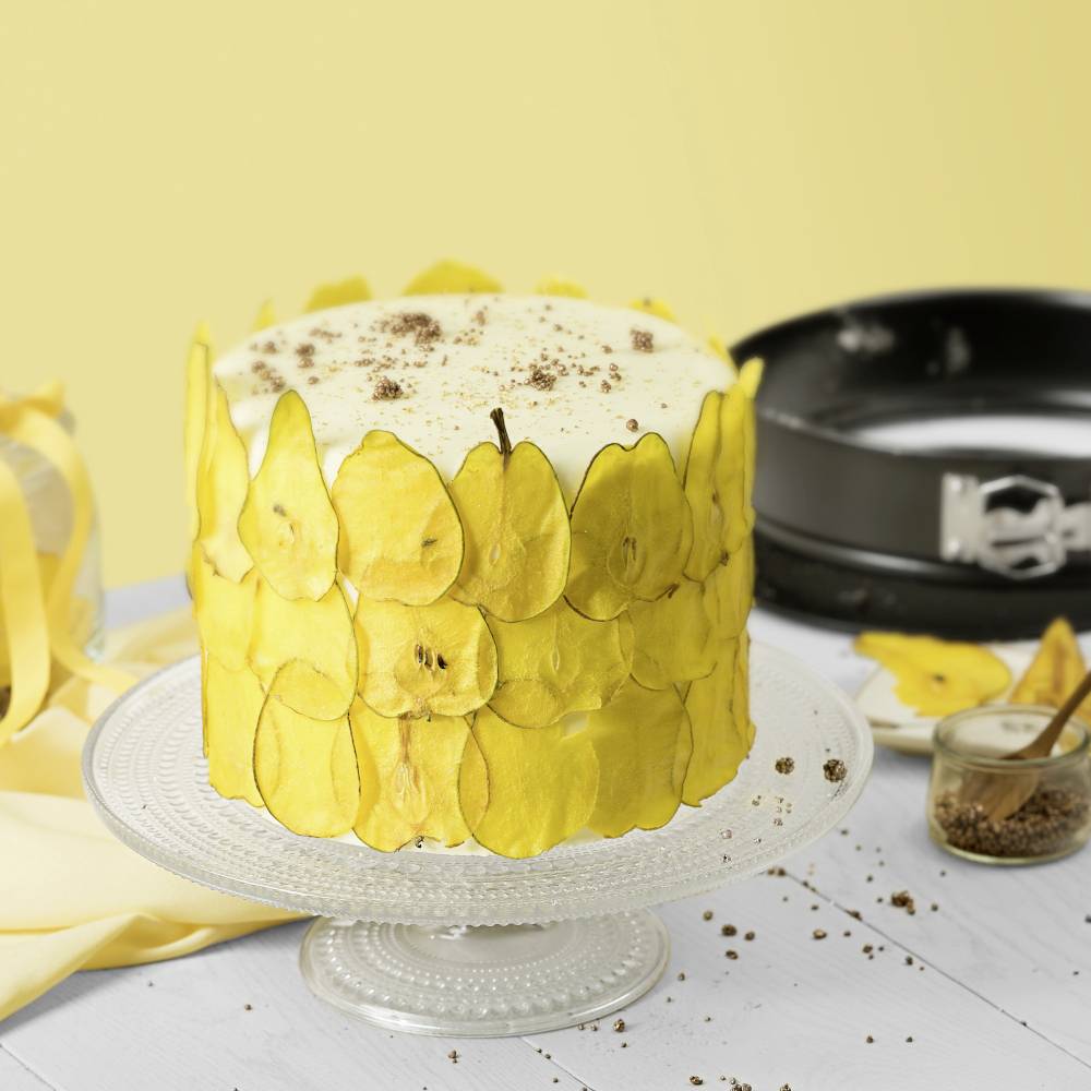 Eine weiß glasierte Torte mit dünnen gelben Fruchtscheiben dekoriert, steht auf einem Glasständer. Daneben eine Backform und Gewürzglas.