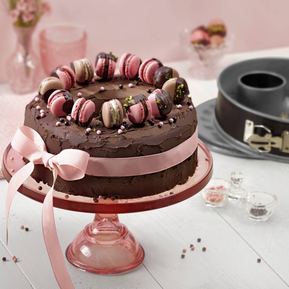 Eine Schokoladentorte mit rosa Schleife auf einem rosa Ständer, dekoriert mit Macarons. Im Hintergrund eine Springform und Gewürze.