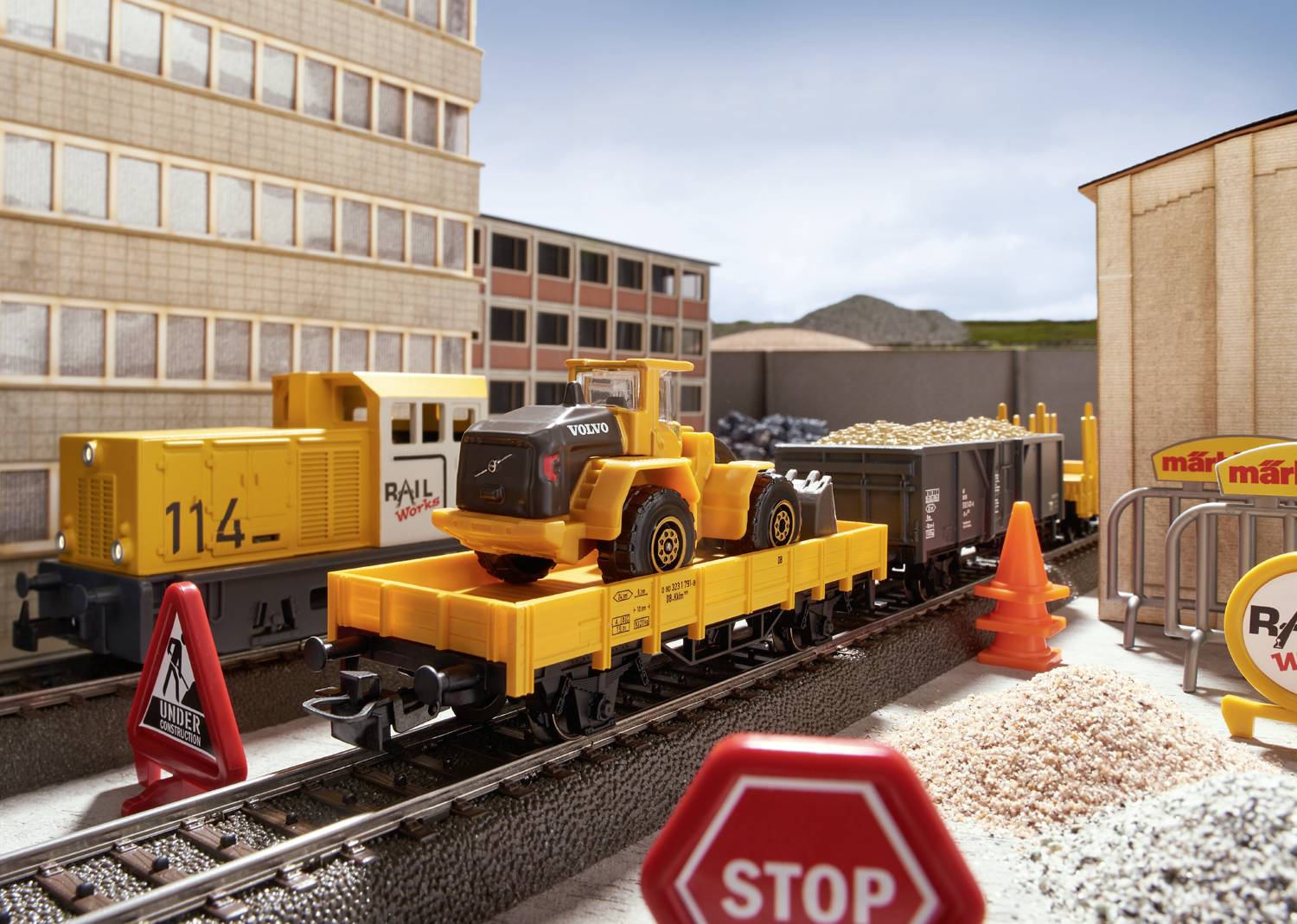 Ein Modellzug mit gelbem Zug und Güterwaggons transportiert eine Spielzeugbaumaschine. Im Hintergrund sind Gebäude zu sehen.