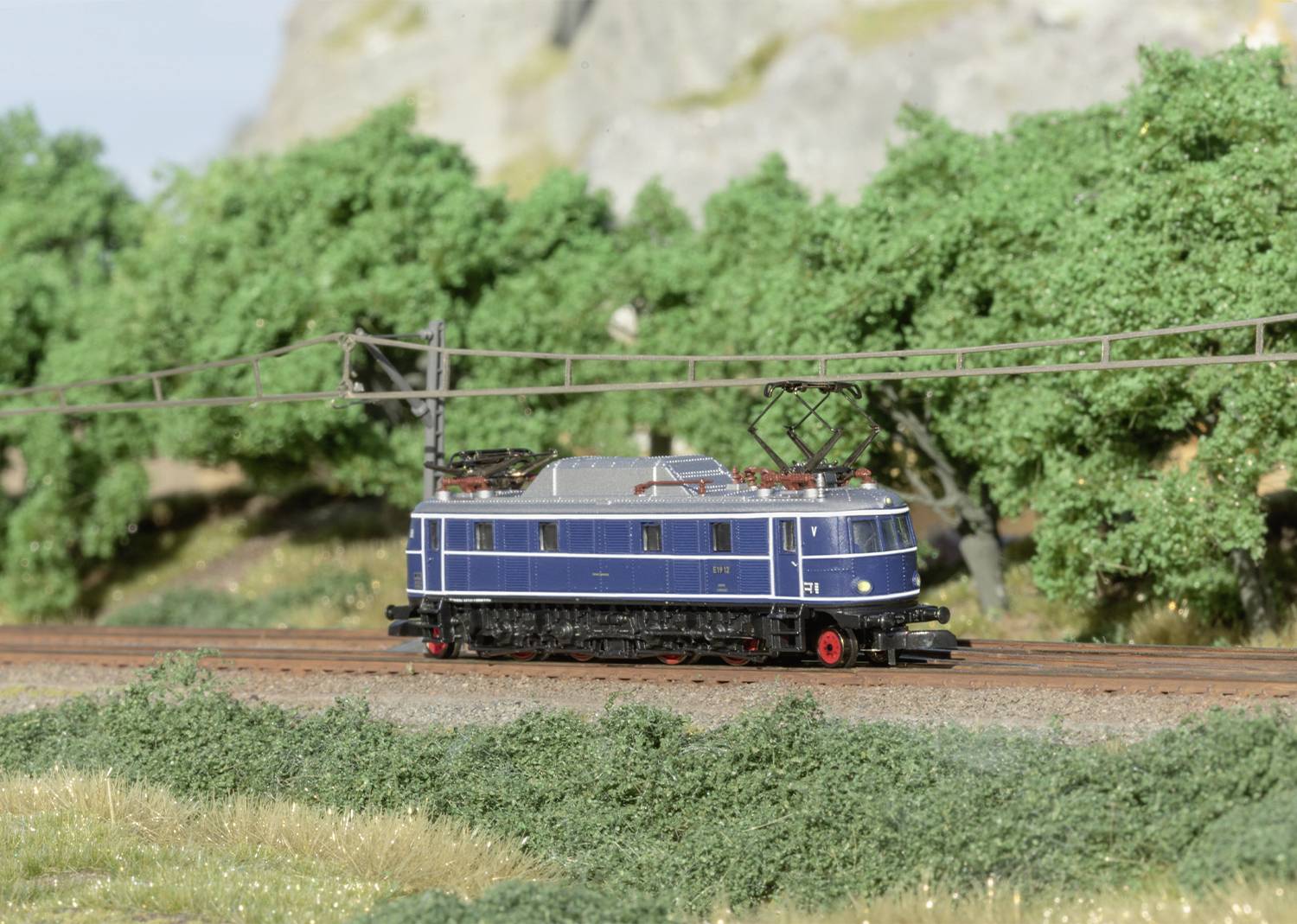 Ein blaues Modellzuglokomotive fährt auf einer Eisenbahnstrecke durch eine grüne Landschaft mit Bäumen im Hintergrund.