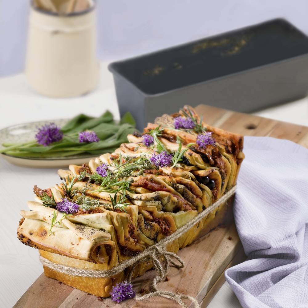 Frisch gebackenes Brot mit Kräutern und violetten Blüten auf einem Holzbrett, daneben ein schwarzer Backbehälter und ein Teller.
