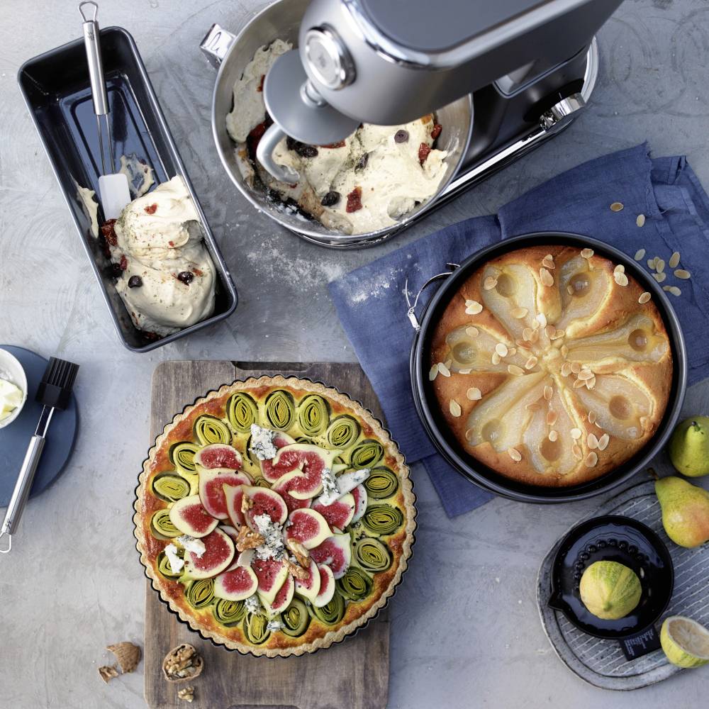 Teigknetmaschine mit Teig und Zutaten. Drei Torten: Feigen-Mandel, Apfel-Mandel und ungefüllte Schale. Dekorativ angerichtete Backzutaten.