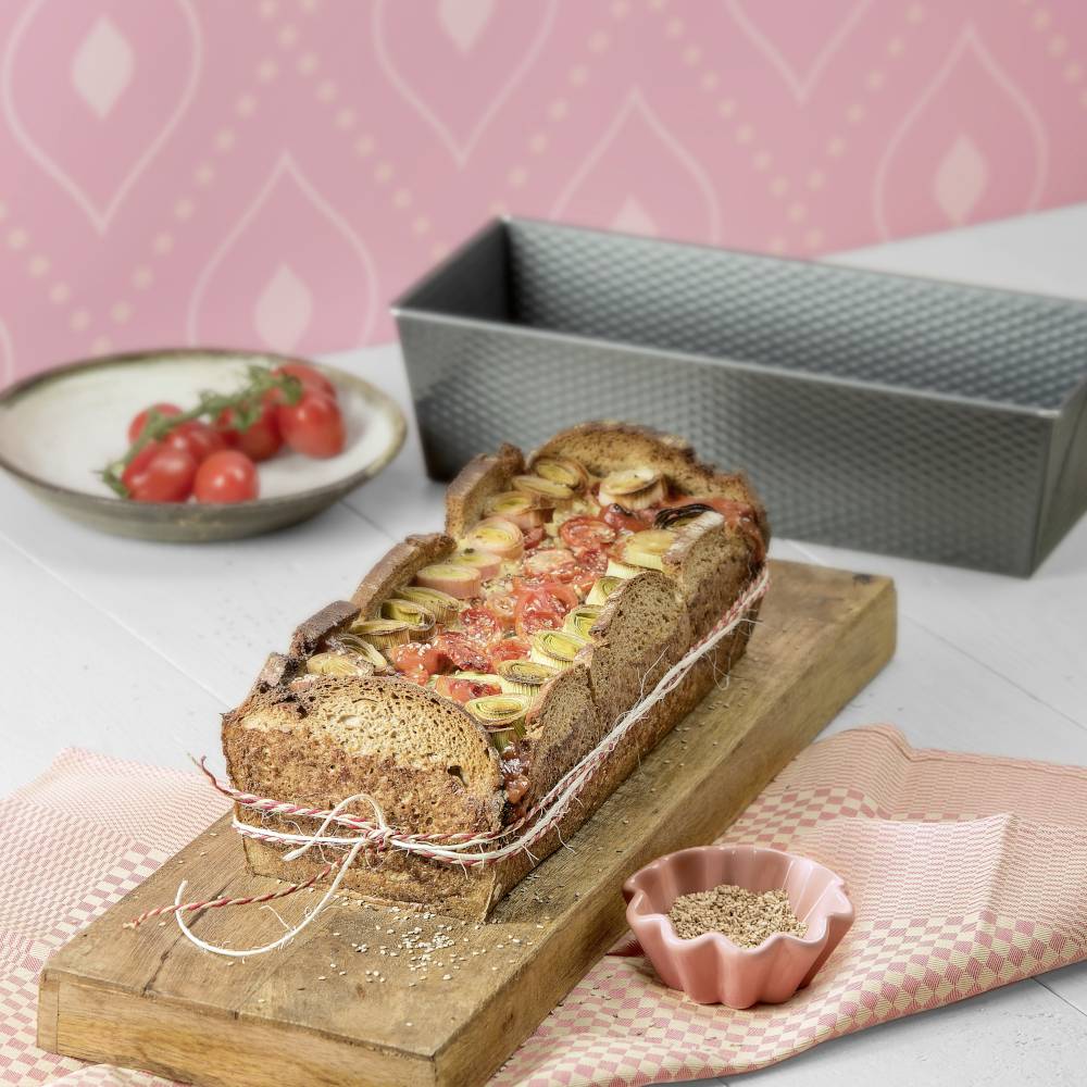 Ein gebackenes Baguette, gefüllt mit Tomaten, Zwiebeln und Kräutern, liegt auf einem Holzbrett. Im Hintergrund eine Schale mit Tomaten.