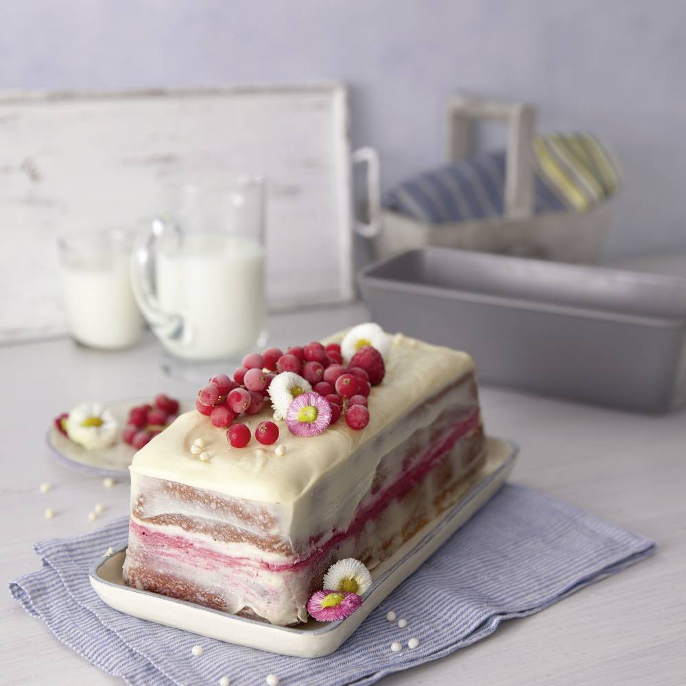 Ein Kuchen mit weißem Zuckerguss und roten Johannisbeeren darauf, steht auf einem Tisch mit einem Glas Milch im Hintergrund.