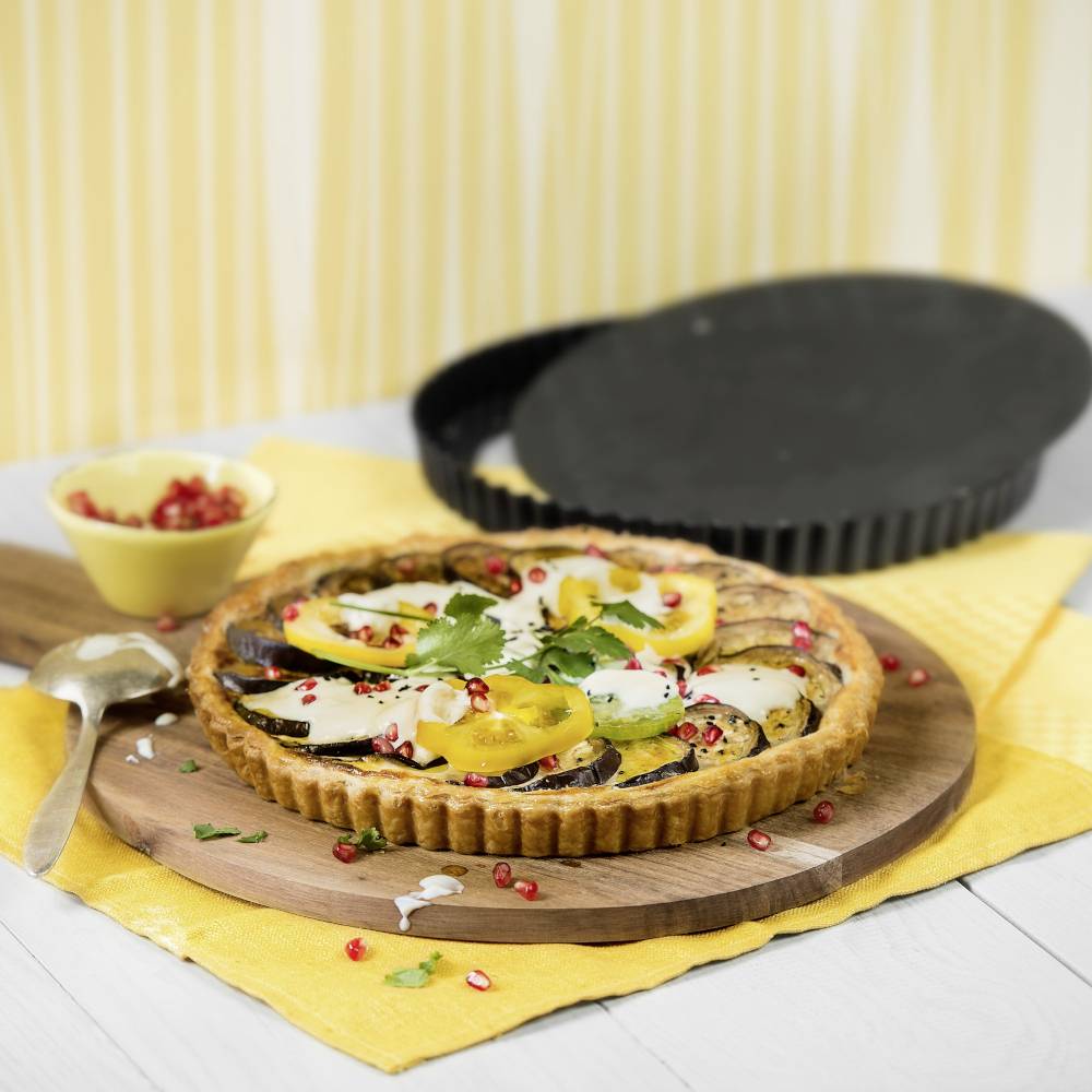 Eine Gemüse-Quiche mit Zucchini, Tomaten und Granatapfelkernen, dekoriert mit Kräutern, auf einem Holzbrett vor gelbem Hintergrund.