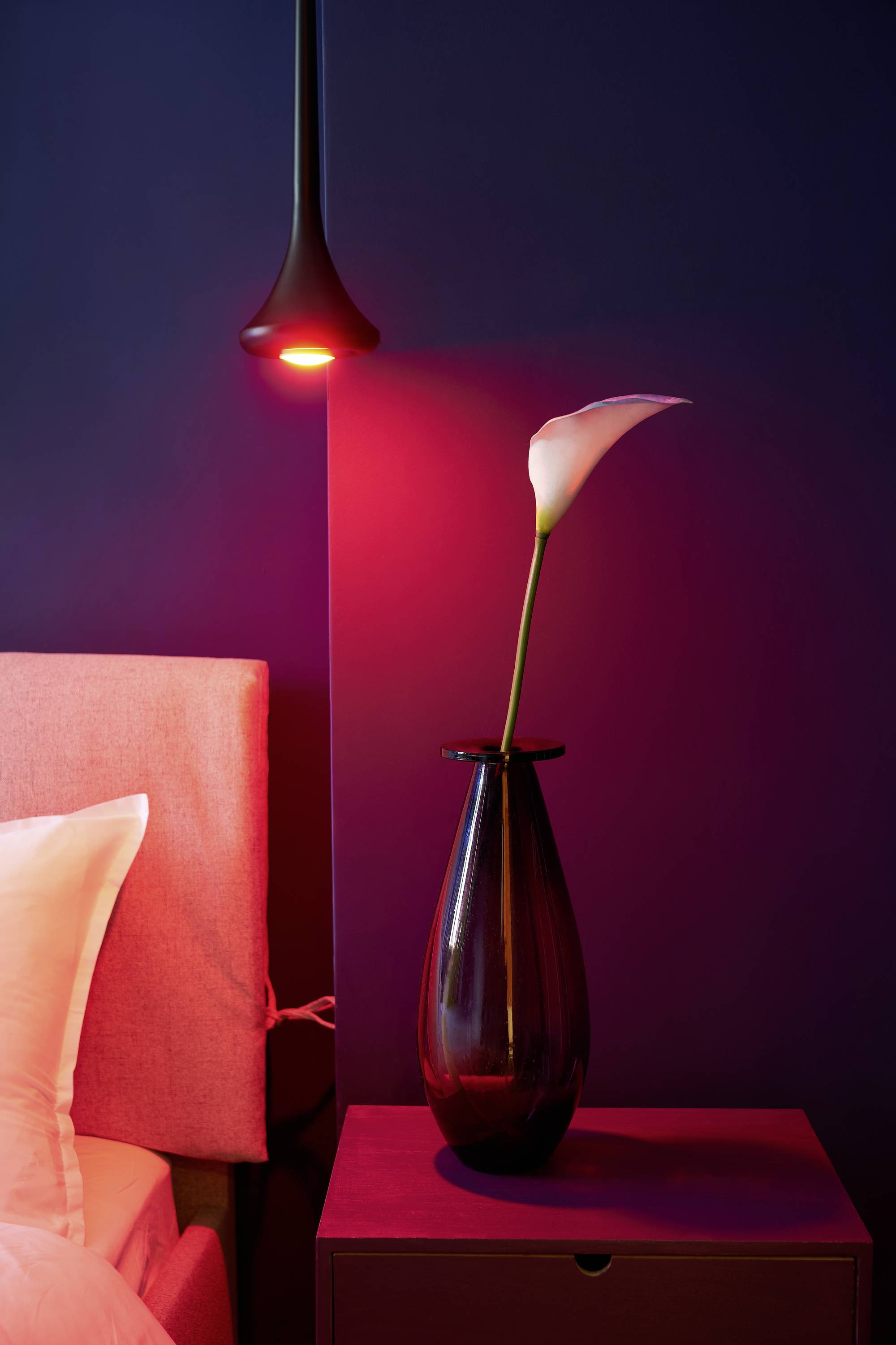 Nachttisch mit einer lilienförmigen Blume in einer dunklen Vase, neben einem Bett. Rotes Licht beleuchtet die Szene von oben.