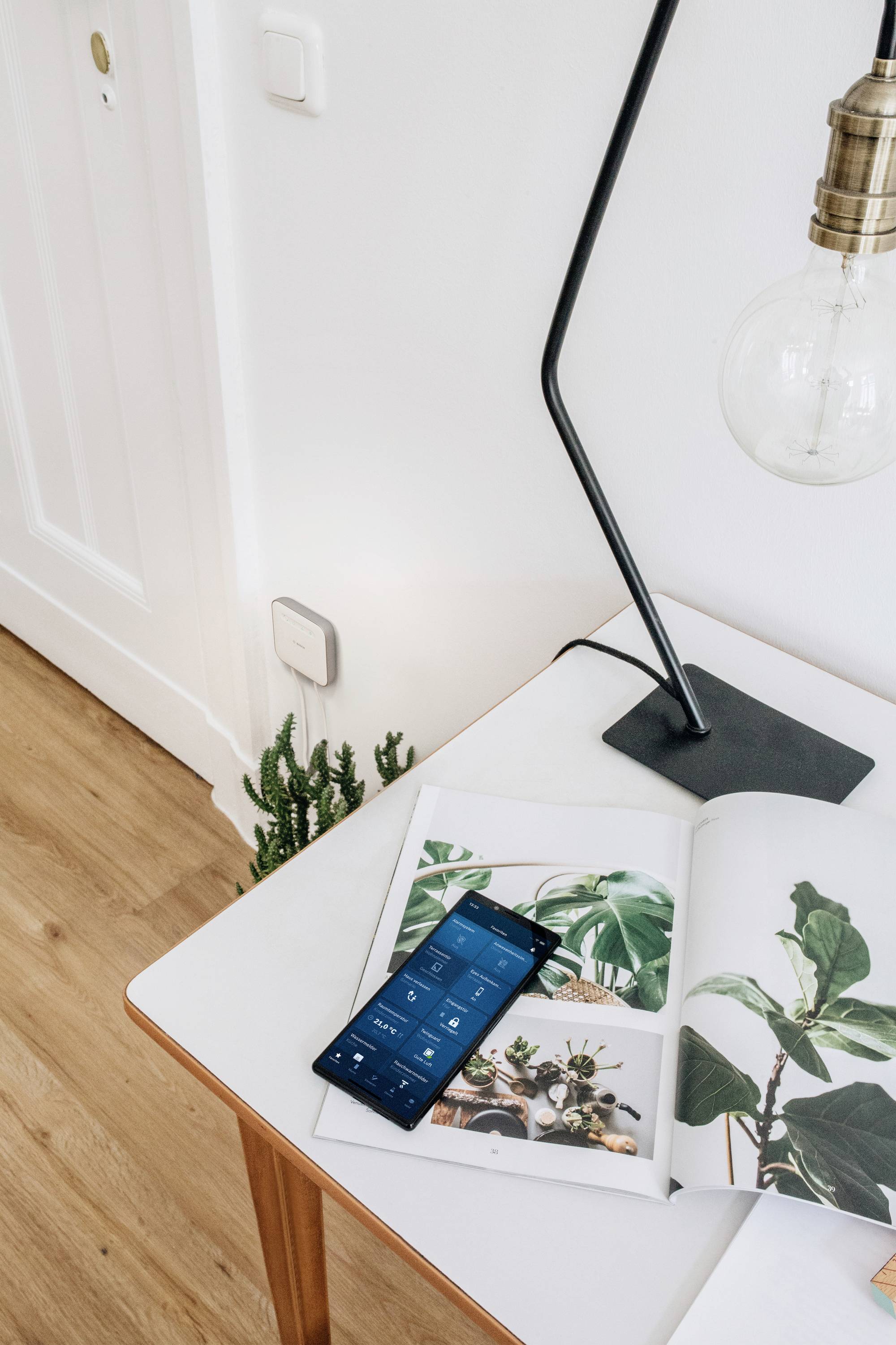 Ein Smartphone liegt auf einem Tisch neben einer geöffneten Zeitschrift mit Pflanzenbildern und einer Lampe.