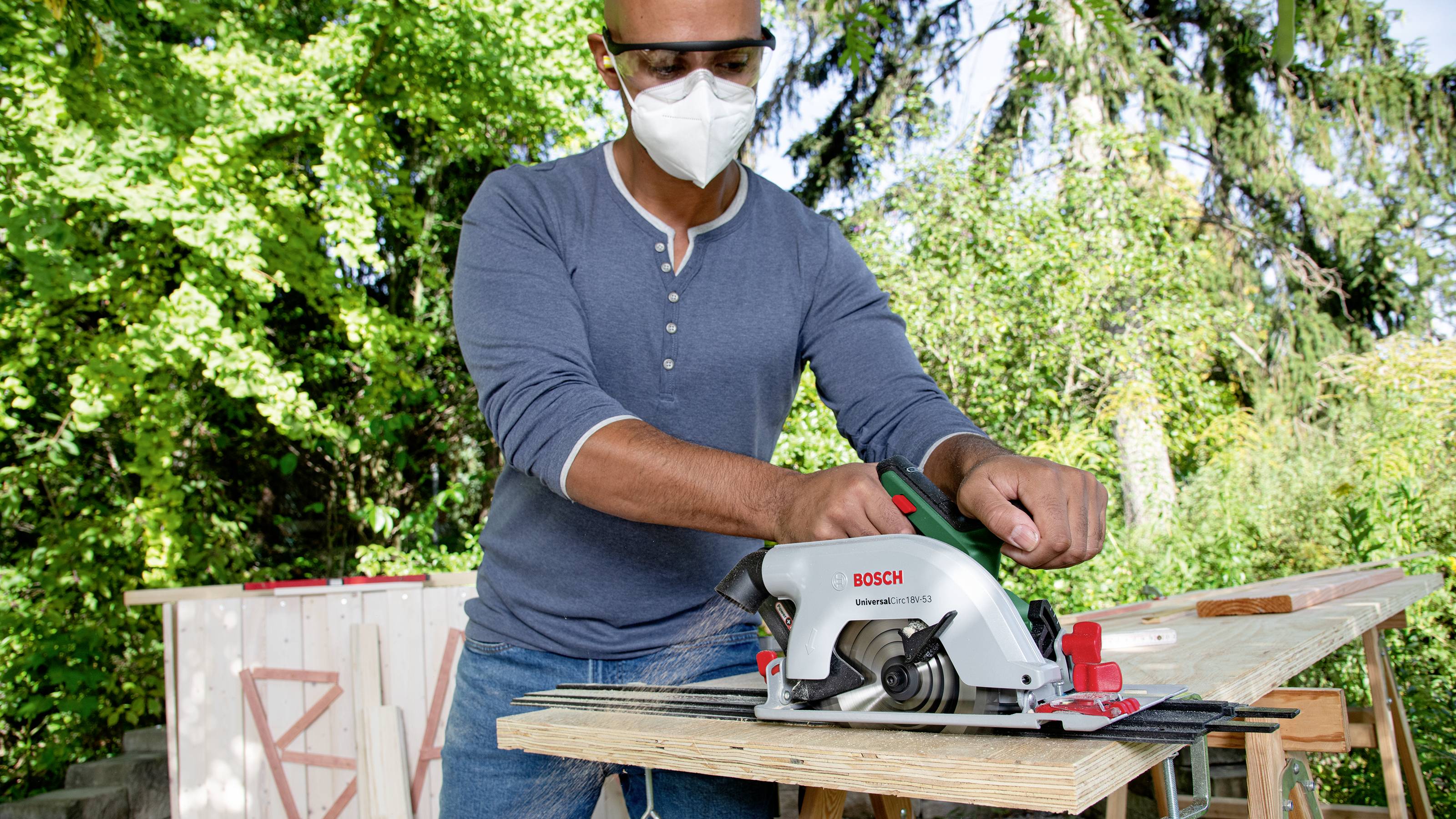 Ein Mann mit Schutzbrille und Maske bearbeitet ein Holzbrett im Freien mit einer Handkreissäge.