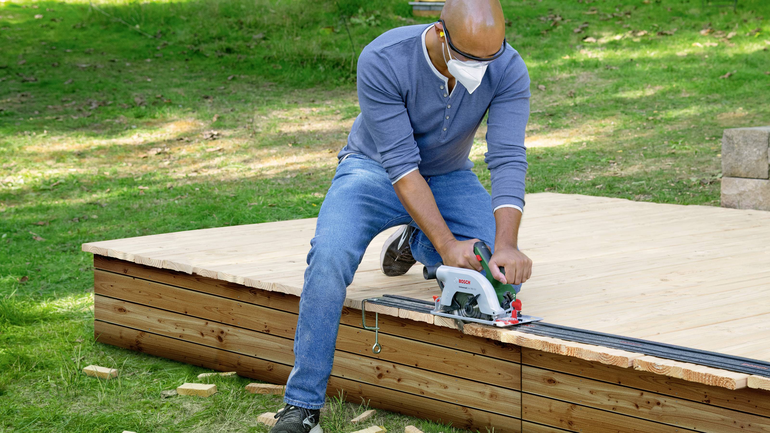 Person sägt Holzterrasse im Garten mit Kreissäge. Trägt Schutzbrille und Maske. Umgebender Rasen und Sonnenschein.